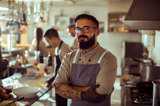 Male restaurant owner with glasses and beard smiling with arms crossed while standing in an industrial kitchen