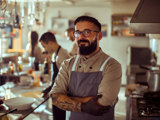 Male restaurant owner with glasses and beard smiling with arms crossed while standing in an industrial kitchen