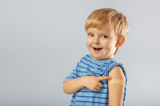 Cute three year old boy in a blue striped shirt smiling to camera pointing to a band-aid after receiving an immunization.