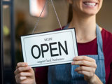 Woman holding 'Open - support local business' sign on the inside door.