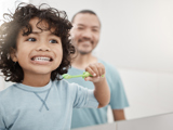 Child smiling while brushing teeth while father watches in the background.